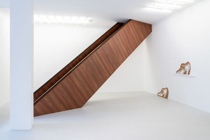 Foto: Einblick in die Ausstellung UHLFELDER im Bildersaal. Eine Ausstellungsansicht in einem weißen, minimalistischen Raum. Im Mittelpunkt steht eine skulpturale Holzkonstruktion in Form einer schrägen Fläche, die an eine Rolltreppe erinnert, mit einer Oberfläche aus Holzfurnier. An der Wand rechts hängen und stehen zwei hellbraune Linolschnitttafeln mit weißen Illustrationen von Schuhen, die wie Schablonen gestaltet sind. Der Raum ist hell beleuchtet, mit weißen Deckenleuchten, die ein modernes Ambiente schaffen.