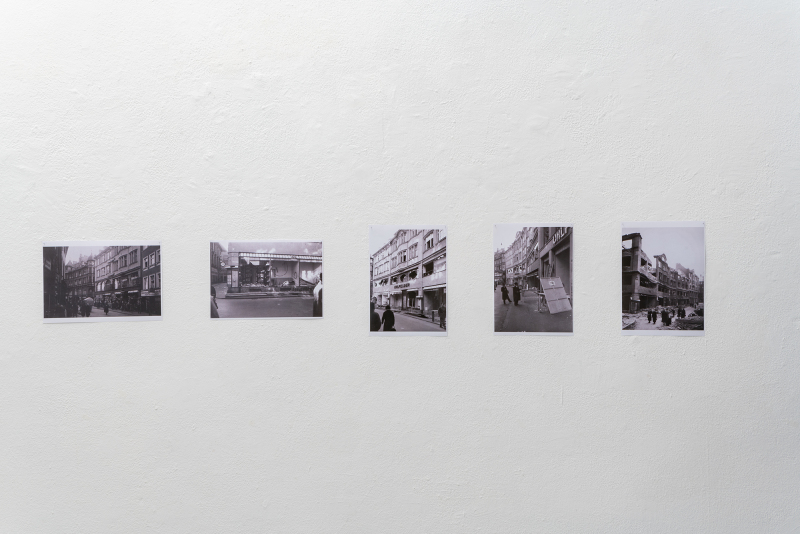 Foto: Nahansicht von Fotografien aus der Ausstellung UHLFELDER. Fünf Schwarz-Weiß-Fotografien sind in einer Reihe an einer weißen Wand angebracht. Die Bilder zeigen verschiedene historische Ansichten des ehemaligen Kaufhauses Uhlfelder im Rosental, vor und nach der Zerstörung. Die Anordnung der Fotos wirkt schlicht und konzentriert sich auf die Dokumentation und den historischen Kontext.