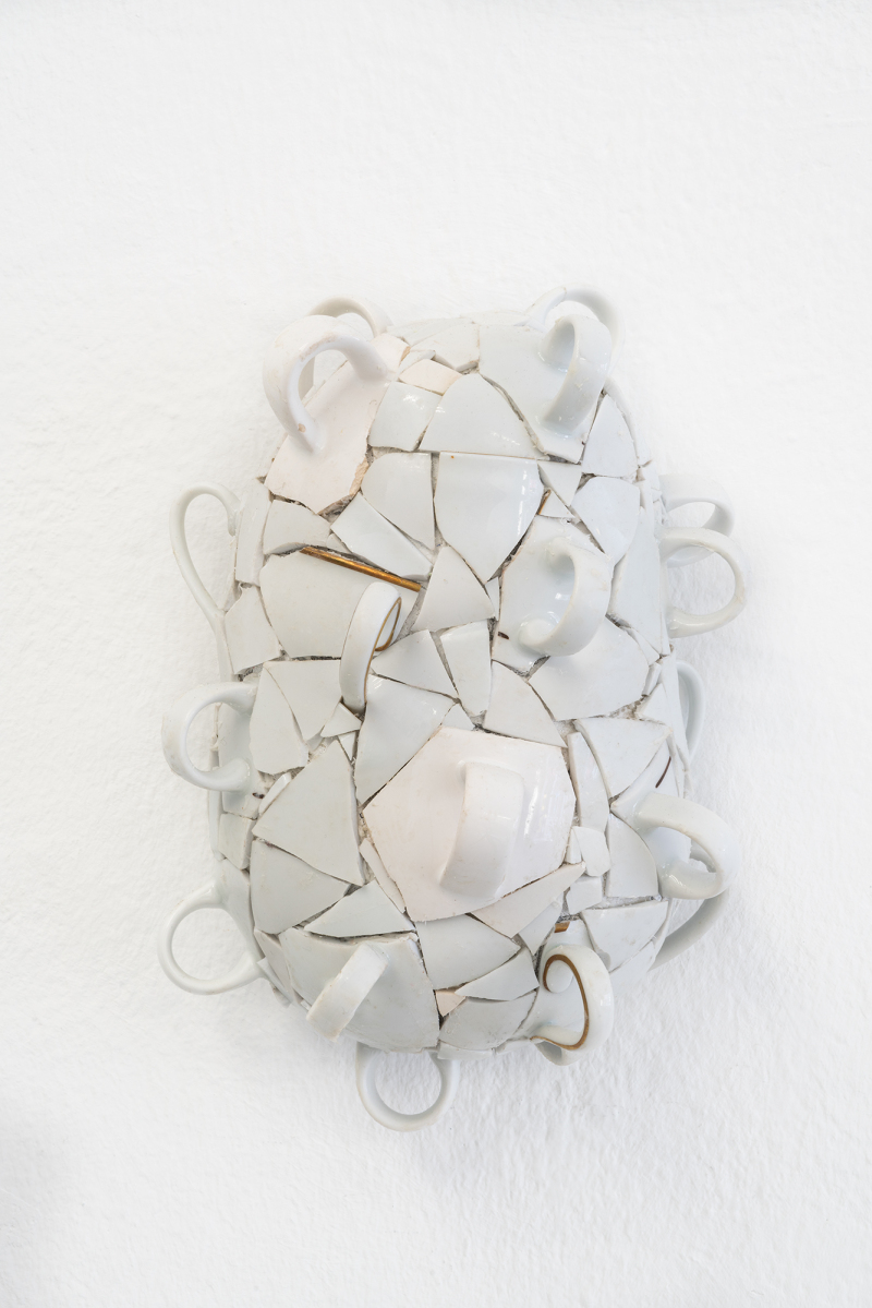 Foto: Detailansicht eines Wandobjekts von Alix Stadtbäumer. Ein Objekt aus weißen, zersprungenen Keramiktassen, das zu einer ovalen, skulpturalen Form zusammengesetzt ist. Die Henkel der Tassen ragen in verschiedene Richtungen heraus, während die Bruchstücke eng aneinandergefügt sind und eine dynamische Struktur erzeugen. Einige der Kanten weisen feine Details wie goldene Akzente auf. Das Kunstwerk ist an einer weißen Wand angebracht und hebt sich durch die komplexe Anordnung der Bruchstücke hervor.